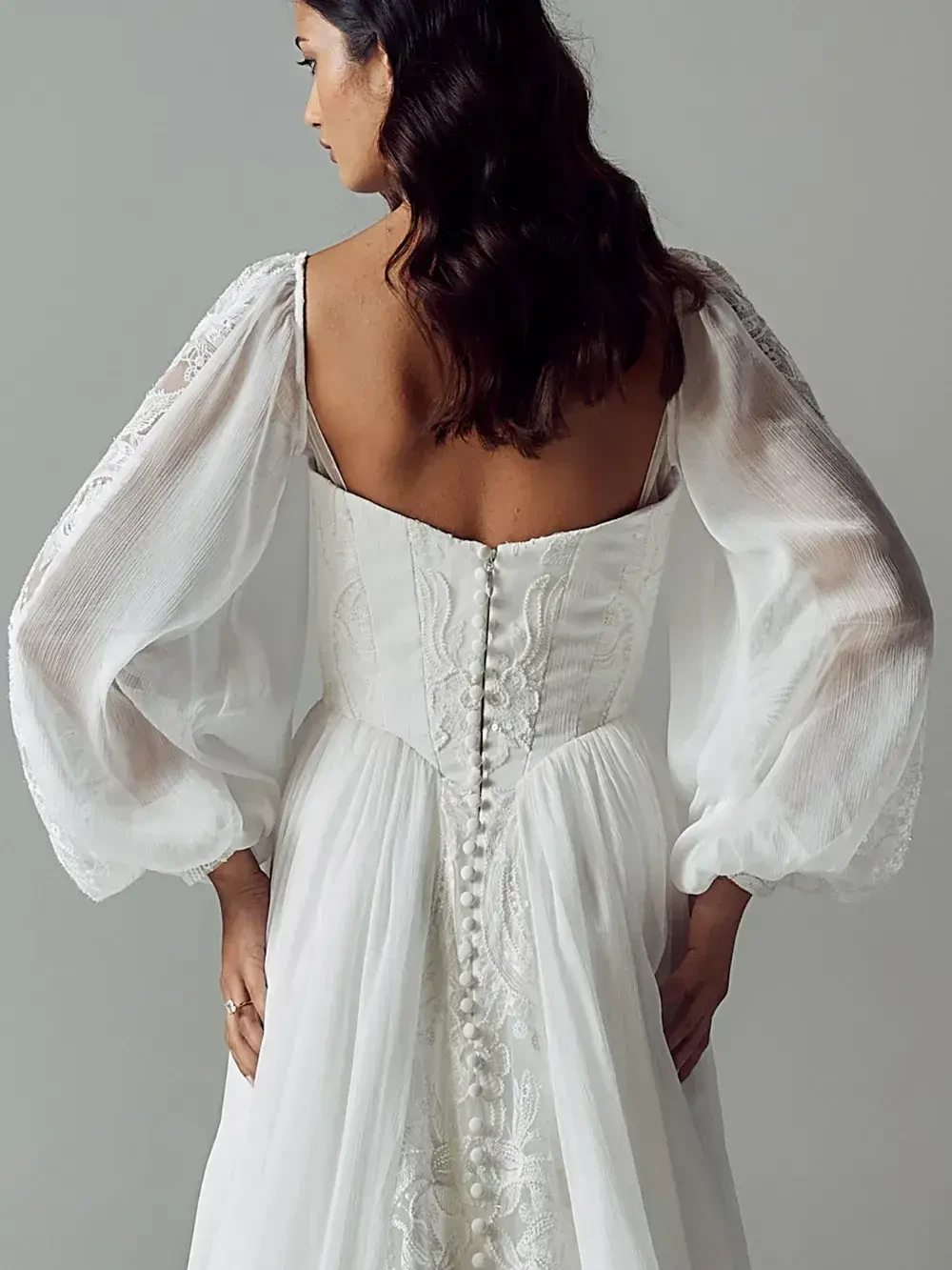 A woman in an elegant white dress with lace detailing and voluminous sleeves stands with her back to the camera. Her wavy hair cascades down.