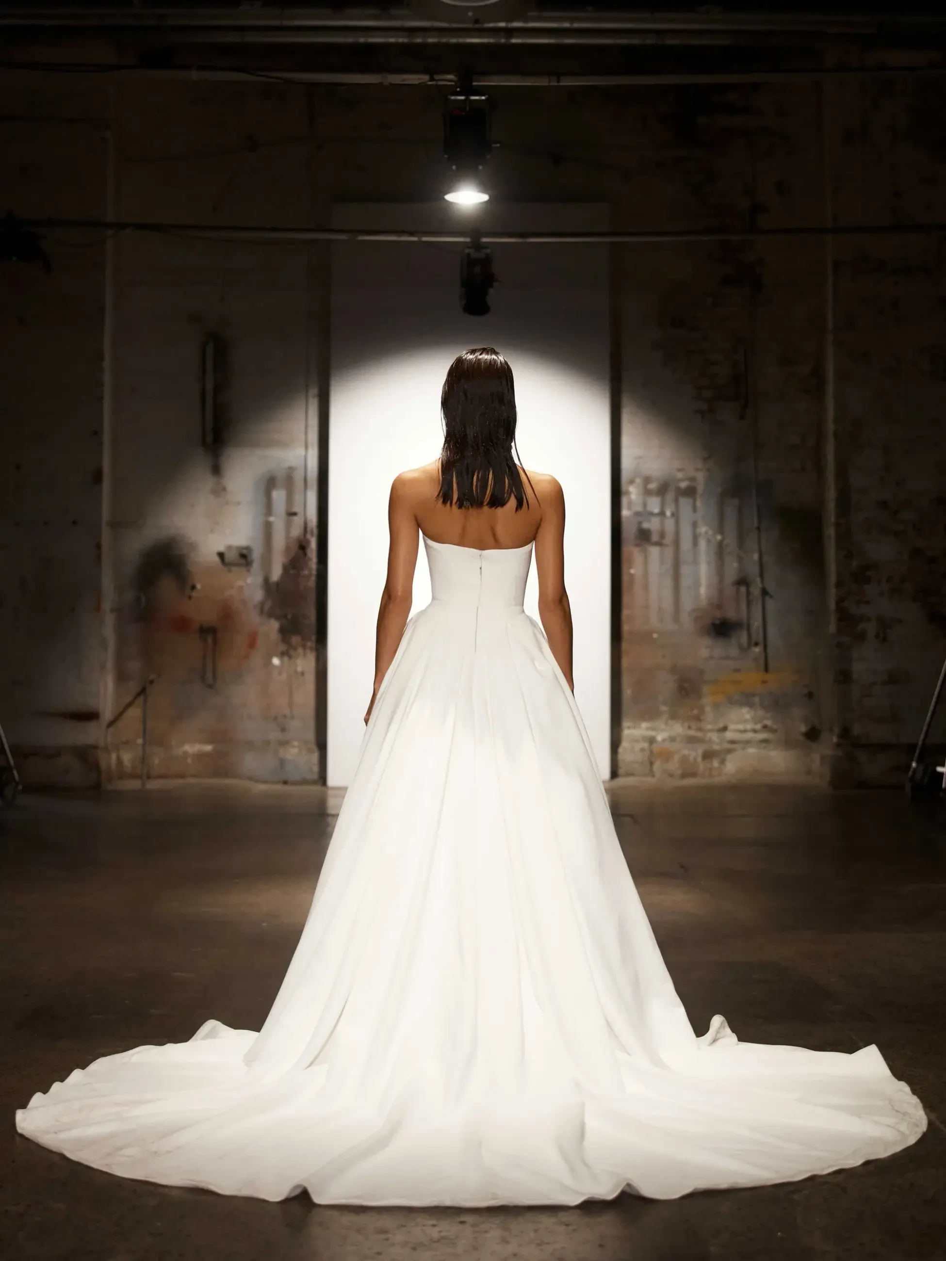A woman in a strapless white gown stands facing a lit backdrop in a dim, industrial space, exuding elegance and contrast between light and shadow.