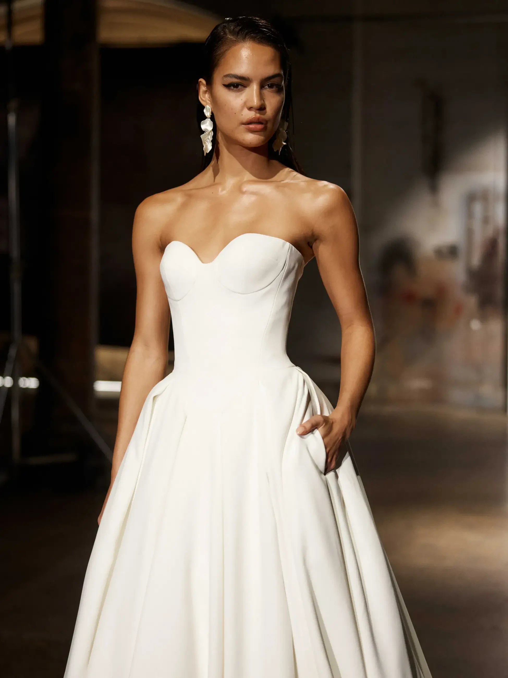 A woman confidently models a strapless white gown with a full skirt in a dimly lit space. She wears statement earrings, conveying elegance and poise.