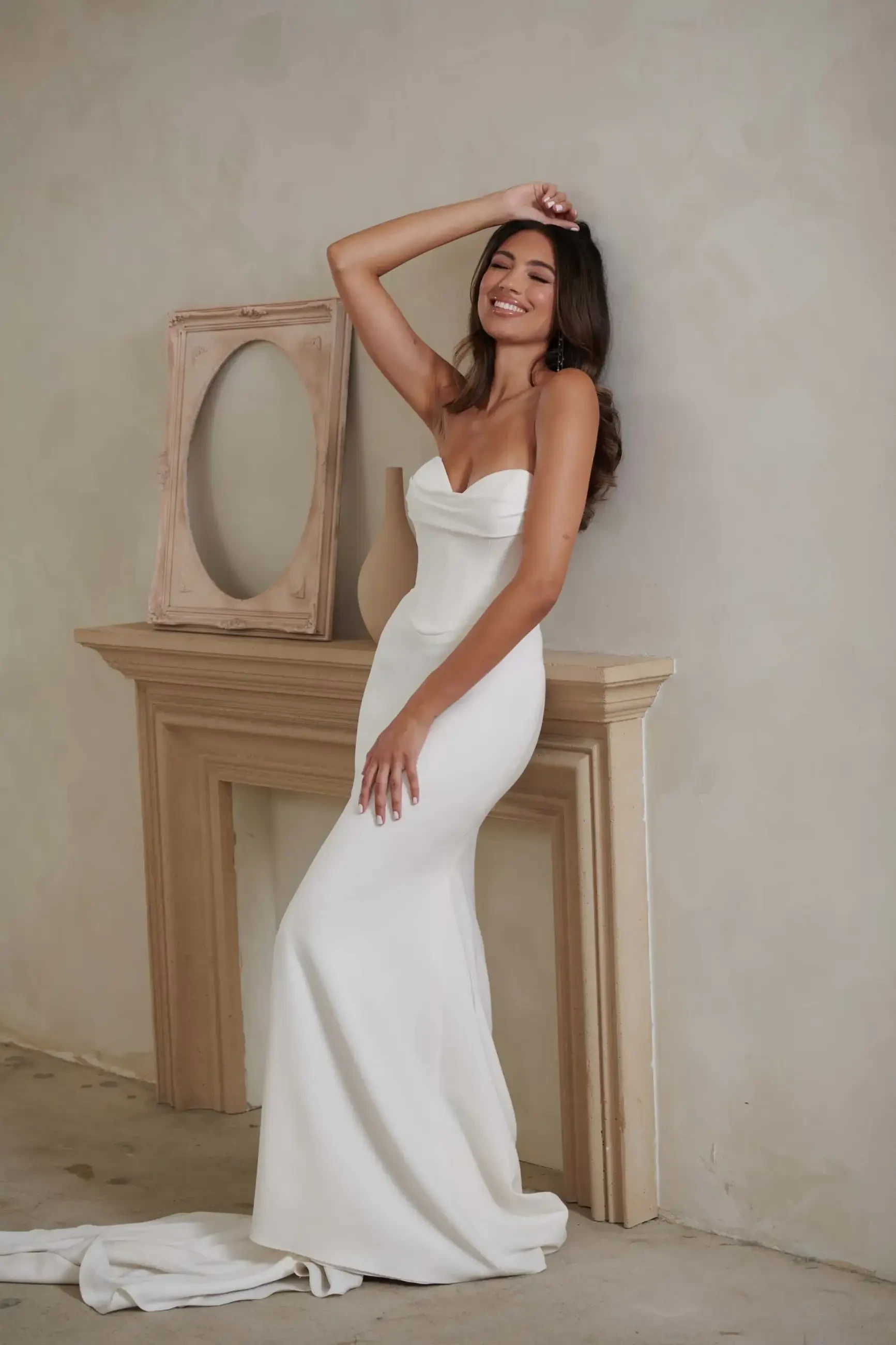 A woman in a strapless white gown leans against a mantel, smiling joyfully. An empty oval frame sits on the mantel, creating an elegant, serene atmosphere.