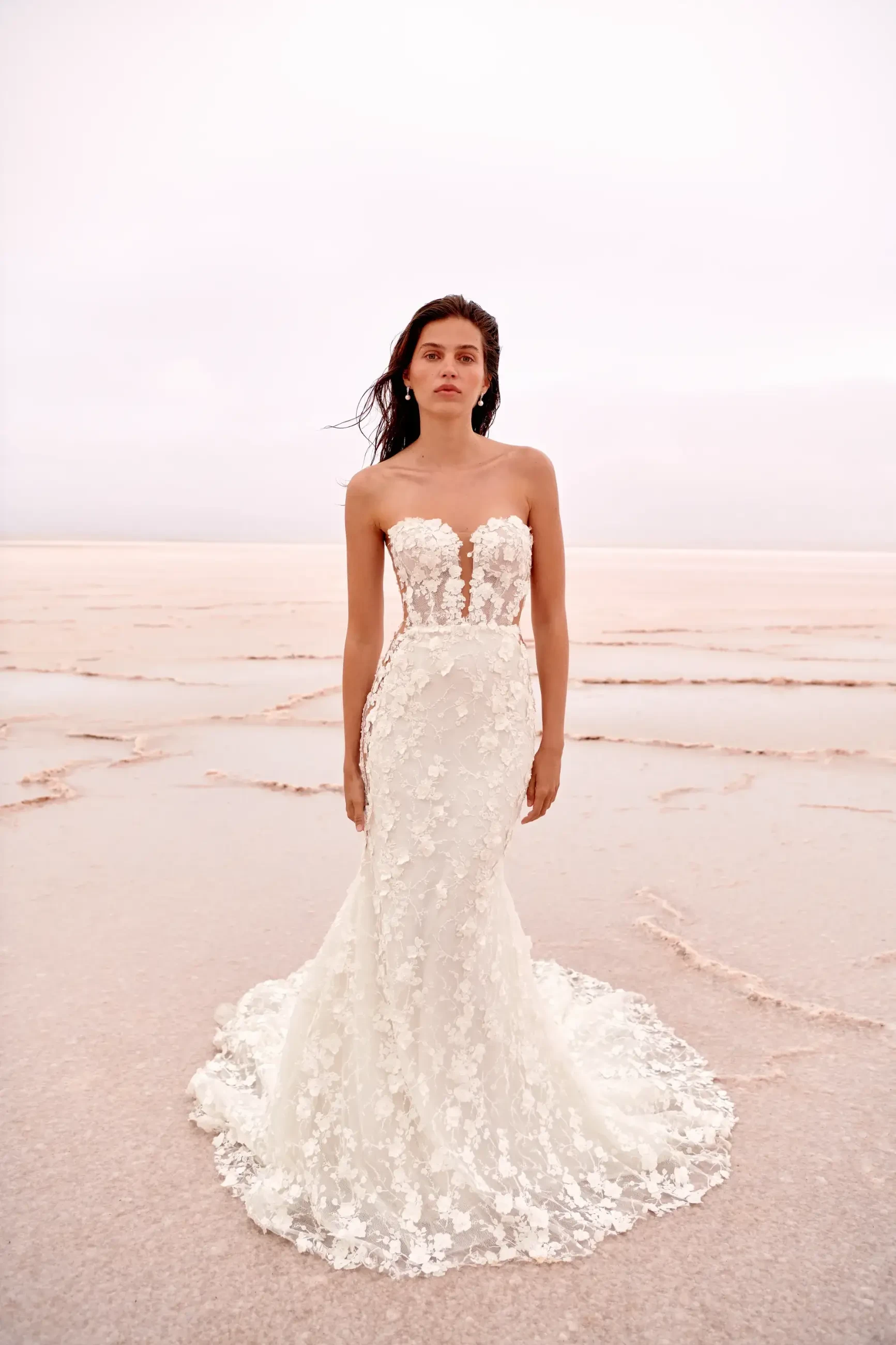 A woman stands on a pale, sandy terrain, wearing a strapless lace bridal gown with floral details. Her expression is serene, set against a soft pink horizon.