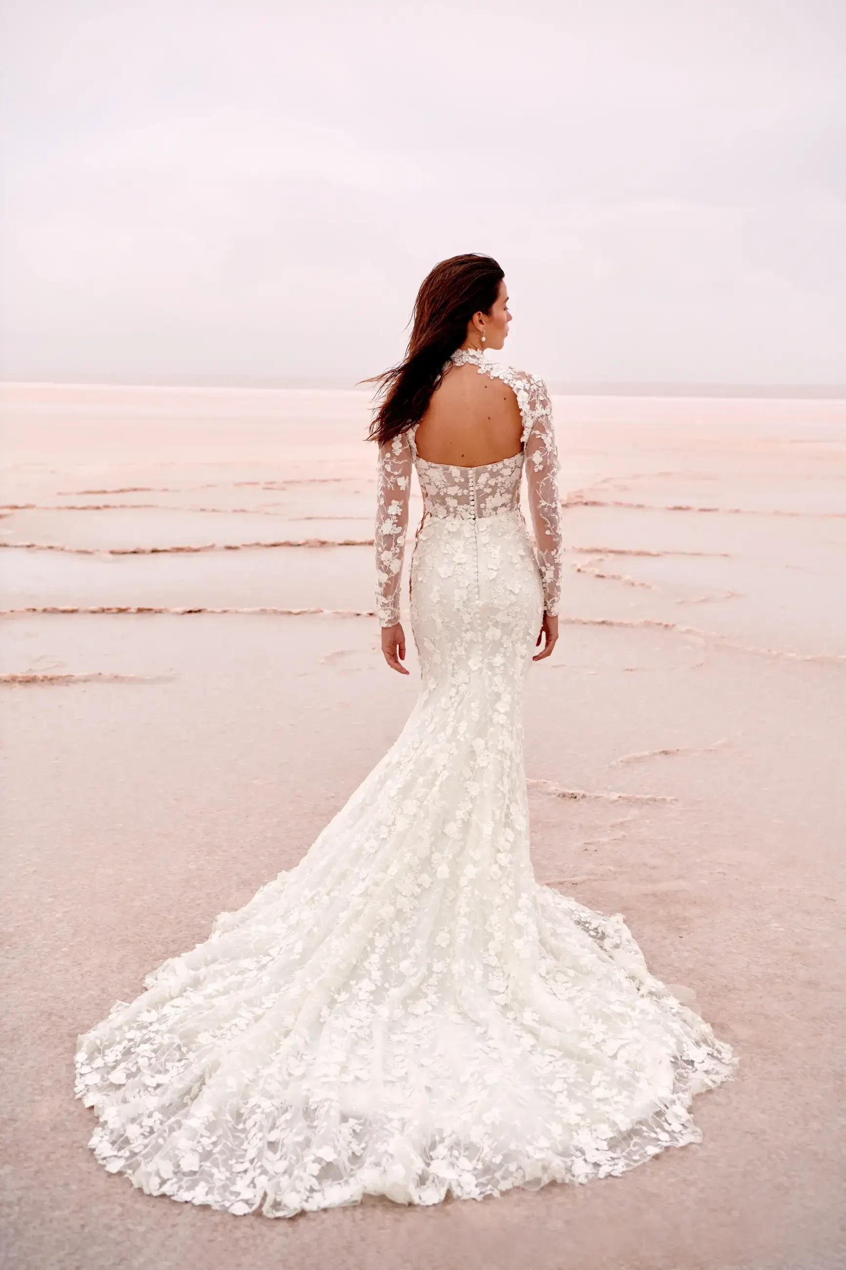 A woman in an elegant lace wedding dress stands on a pink-tinted beach, her back facing the camera. The scene feels serene and romantic.