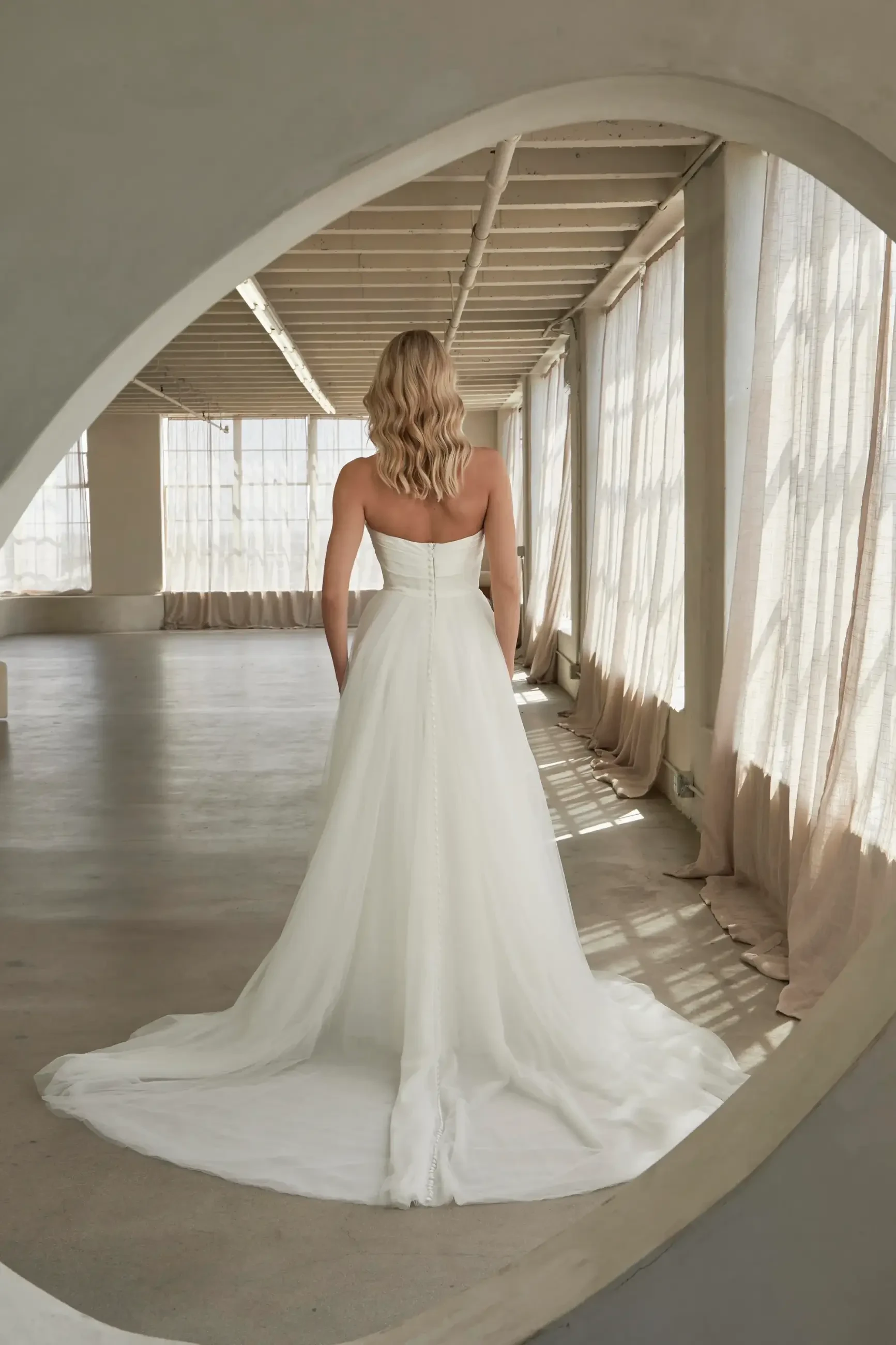 A woman in a flowing white strapless wedding gown stands with her back turned in a sunlit, airy room with tall windows and light curtains.