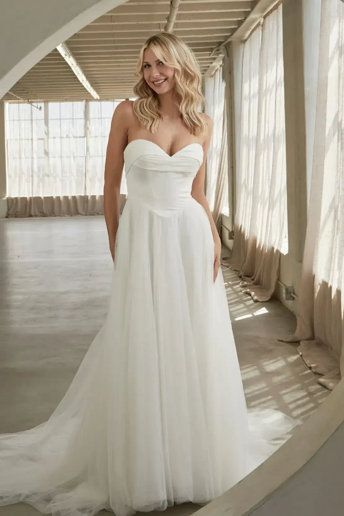 A woman in a flowing white strapless wedding dress stands in a sunlit room with sheer curtains. She smiles gently, conveying joy and elegance.