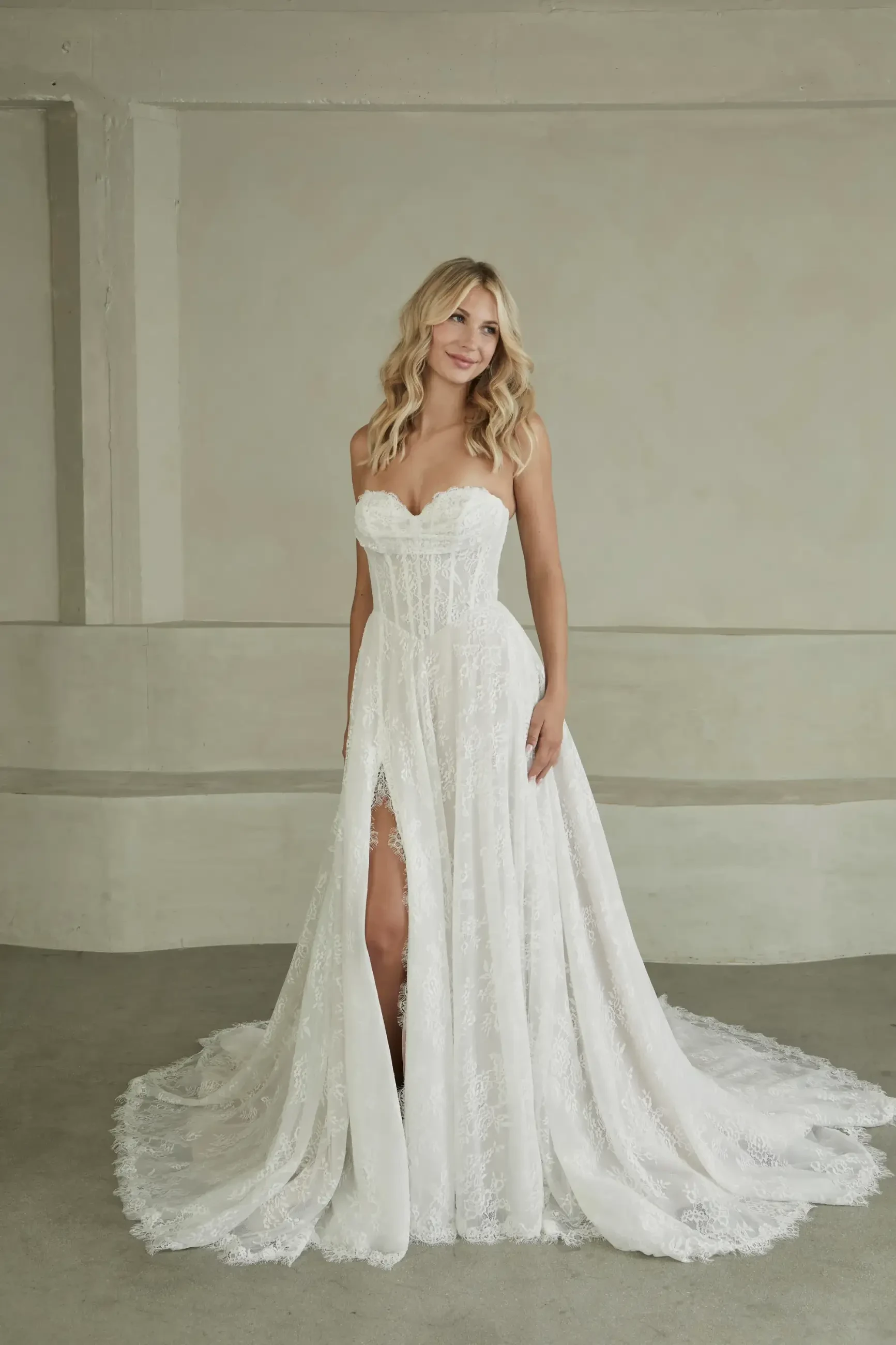 Blonde woman in an elegant, strapless lace wedding gown with a sweetheart neckline and thigh-high slit. She smiles softly in a minimalistic setting.