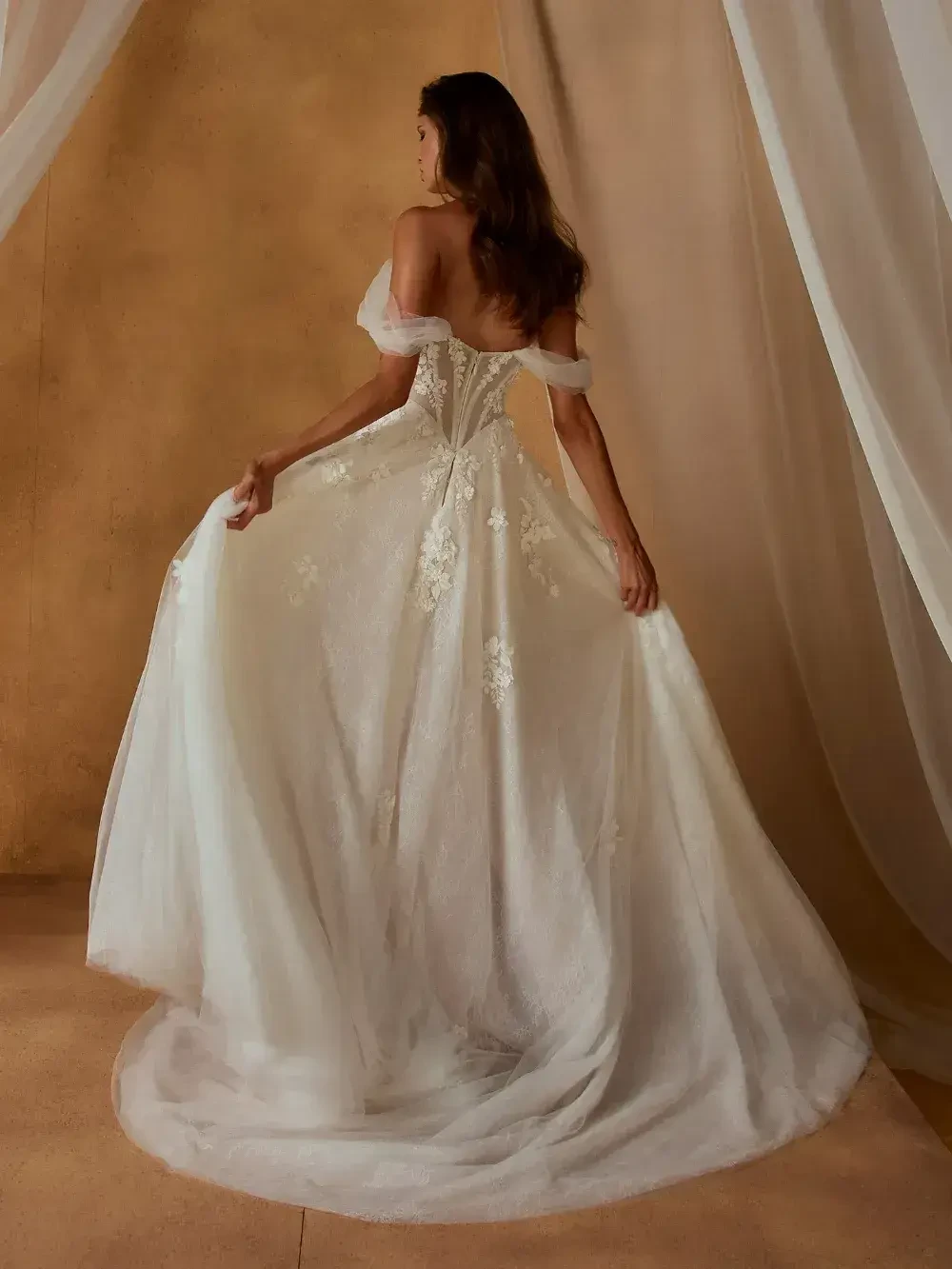 A woman is standing in an elegant off-shoulder wedding dress with floral lace details, holding the flowing skirt. The background features soft, draped fabric and a warm beige wall, conveying a romantic and serene atmosphere.