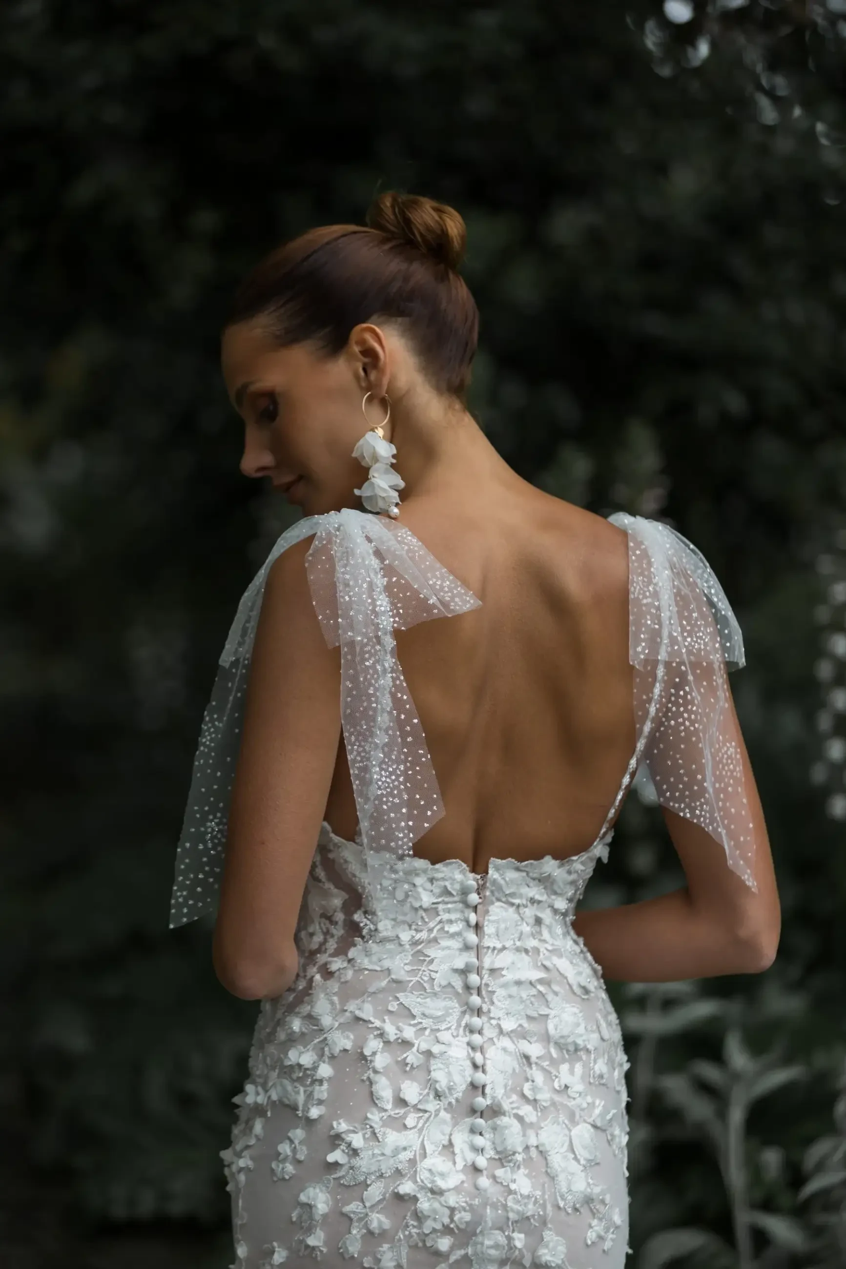 Woman in an elegant backless wedding dress with floral lace and sheer straps, gazing down. Her hair is in a bun, and she wears large floral earrings.
