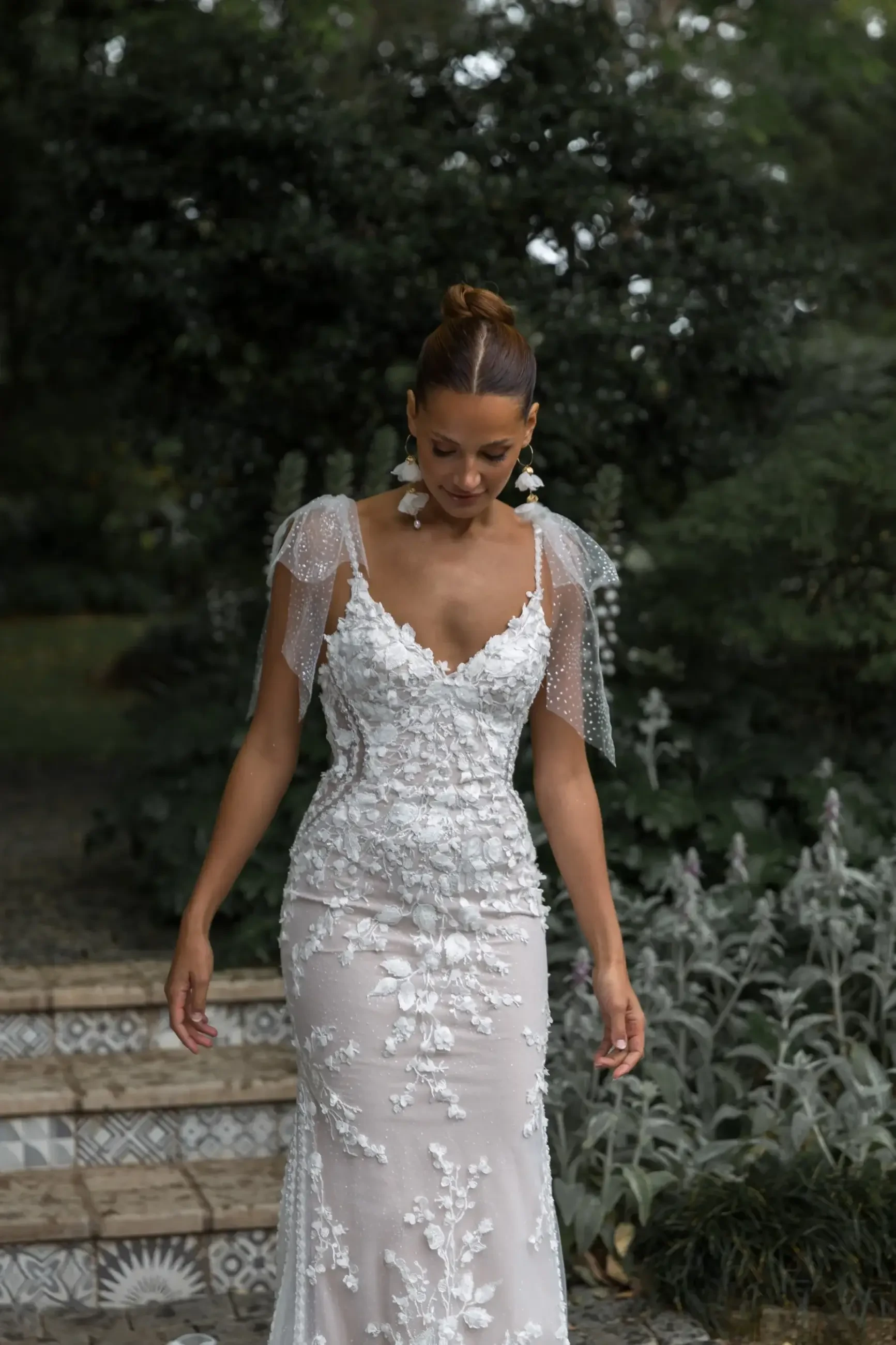 A woman in a detailed lace wedding dress with floral patterns walks down decorated steps. Her hair is in an updo, and the background is a lush garden.