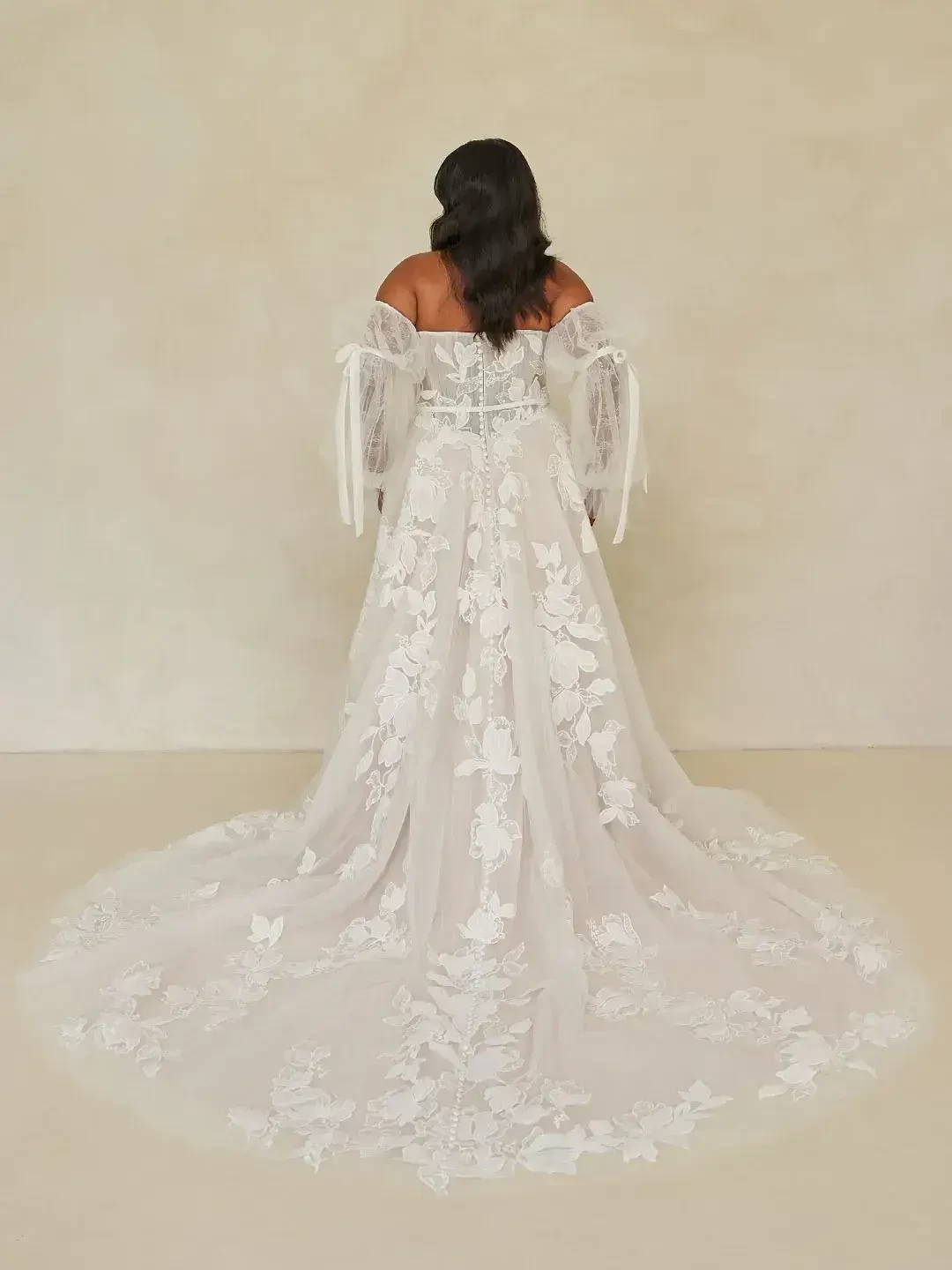Back view of a bride in an elegant off-shoulder gown with sheer sleeves and intricate floral patterns. The long train flows gracefully on a neutral backdrop.