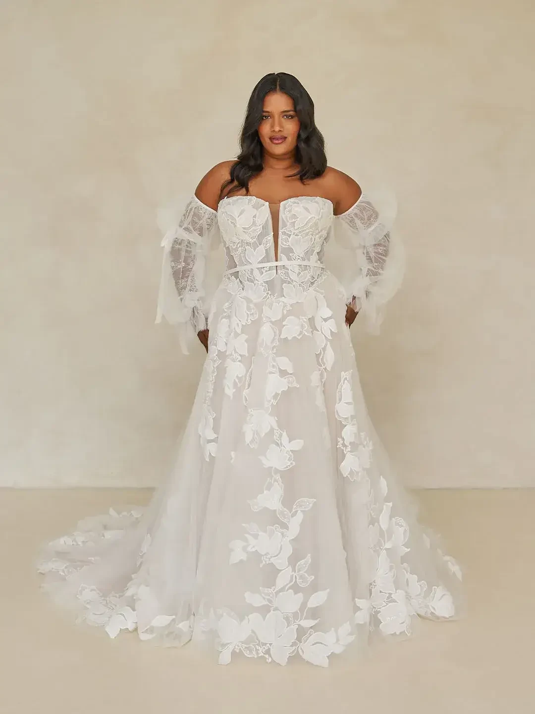 Woman in an elegant, white, off-shoulder wedding gown with floral lace details stands confidently. The sheer, flowing fabric adds a romantic touch.