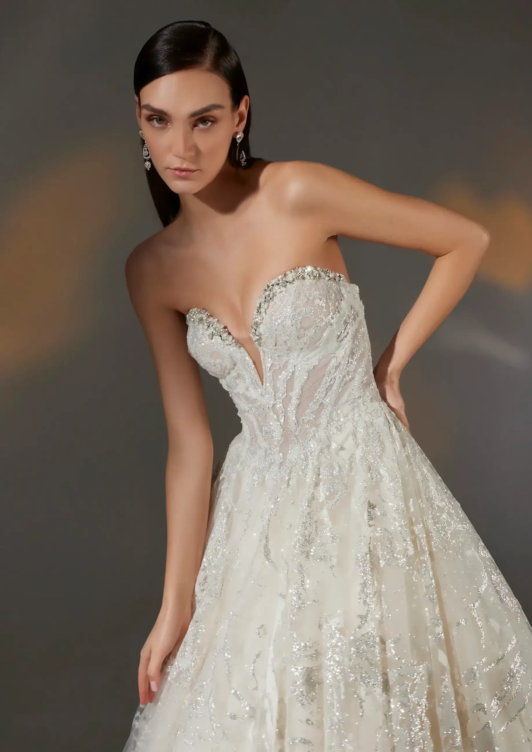 A model wearing a strapless, embellished wedding gown with a deep V-neckline, posed elegantly against a softly lit background.