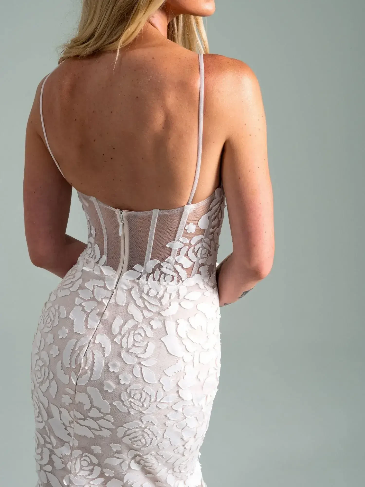 A woman with blonde hair is wearing a backless, form-fitting dress with a floral lace design, evoking elegance and sophistication against a soft gray backdrop.