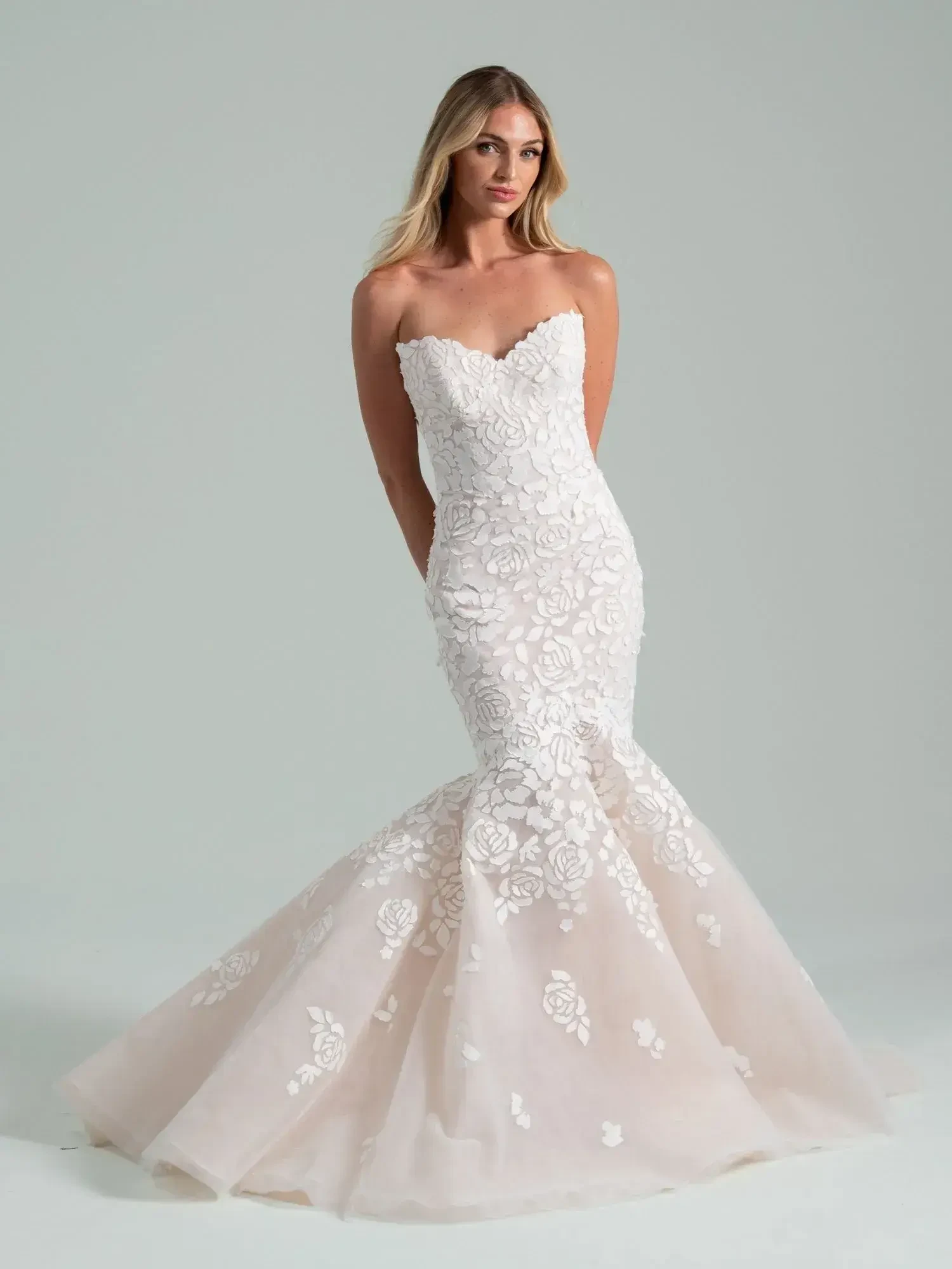 A woman models a strapless, white mermaid-style wedding gown with intricate floral lace patterns, exuding elegance and sophistication against a light gray background.