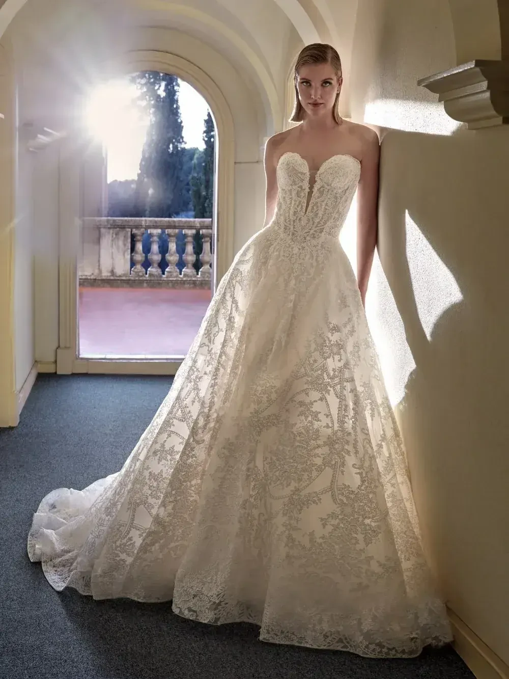 A model in a strapless, intricate lace wedding gown stands beside a wall with sunlight streaming in, casting a soft glow.