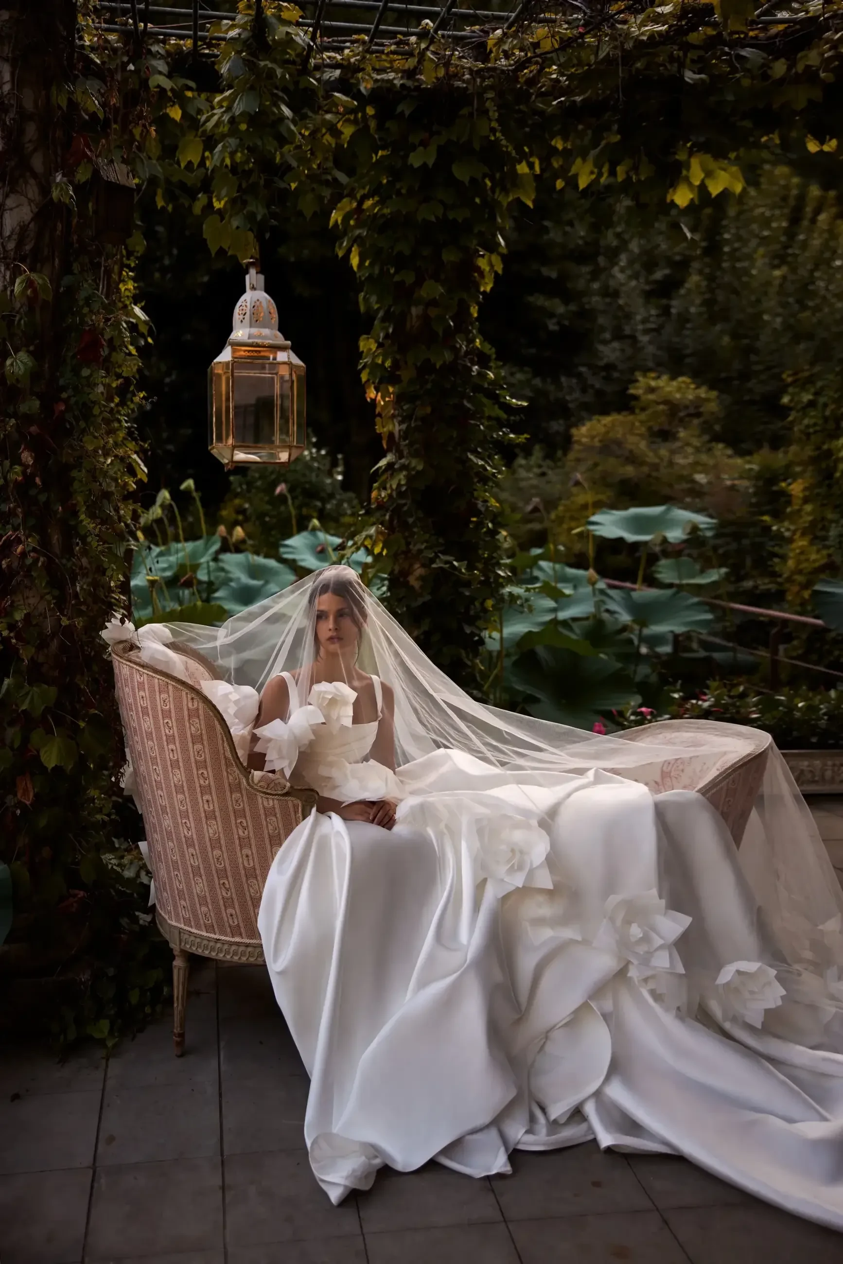 A bride sitting in a beautifully decorated outdoor setting, wearing an elegant wedding gown and veil, surrounded by greenery and soft lighting.