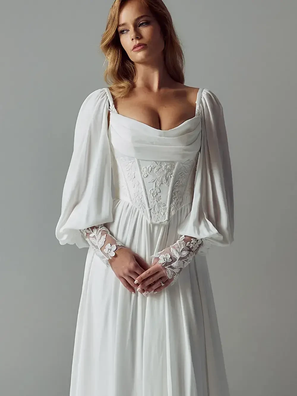 A woman wearing a white dress with puffy sleeves and intricate lace details on the arms, standing against a gray background.