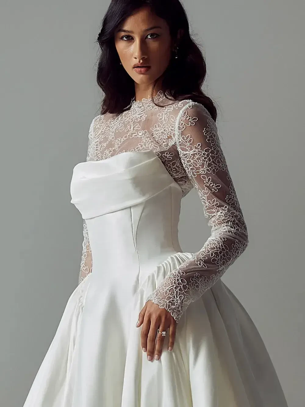 A woman in a white dress with long lace sleeves and a fitted bodice, looking directly at the camera.