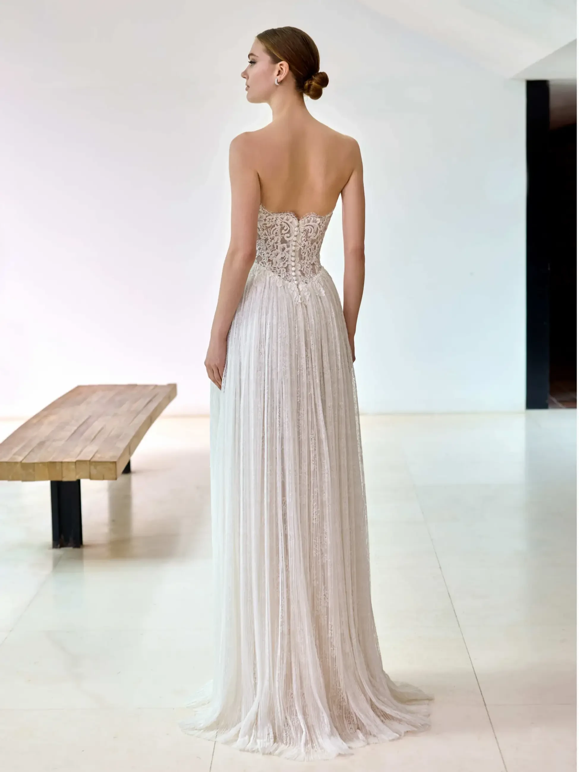 A woman in an elegant, strapless white gown with a lace bodice and pleated skirt stands in a minimalist room, exuding grace and sophistication.