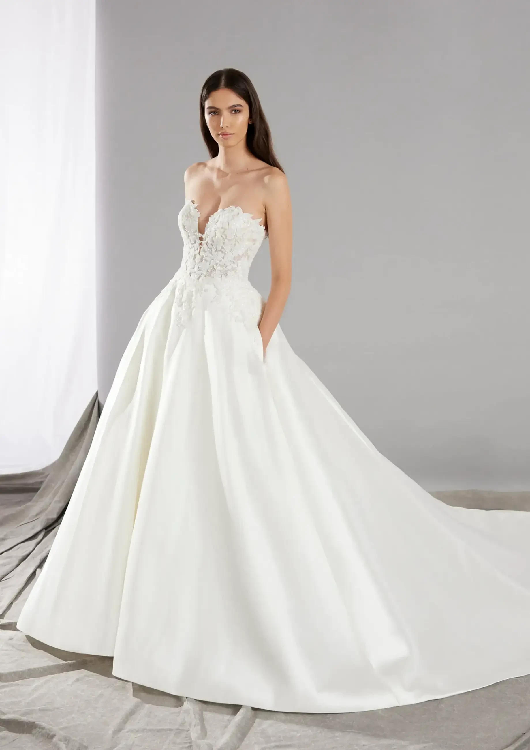 A model is wearing a strapless white wedding gown with a fitted bodice and a full skirt. The gown features delicate floral embroidery and pockets, set against a neutral background.