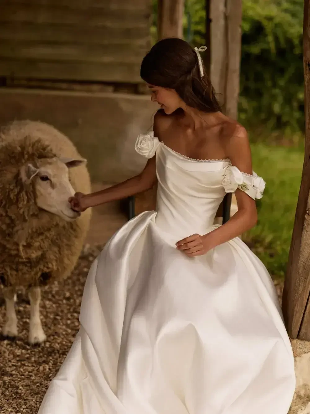 A woman in a white wedding dress interacts with a sheep in a rustic setting.
