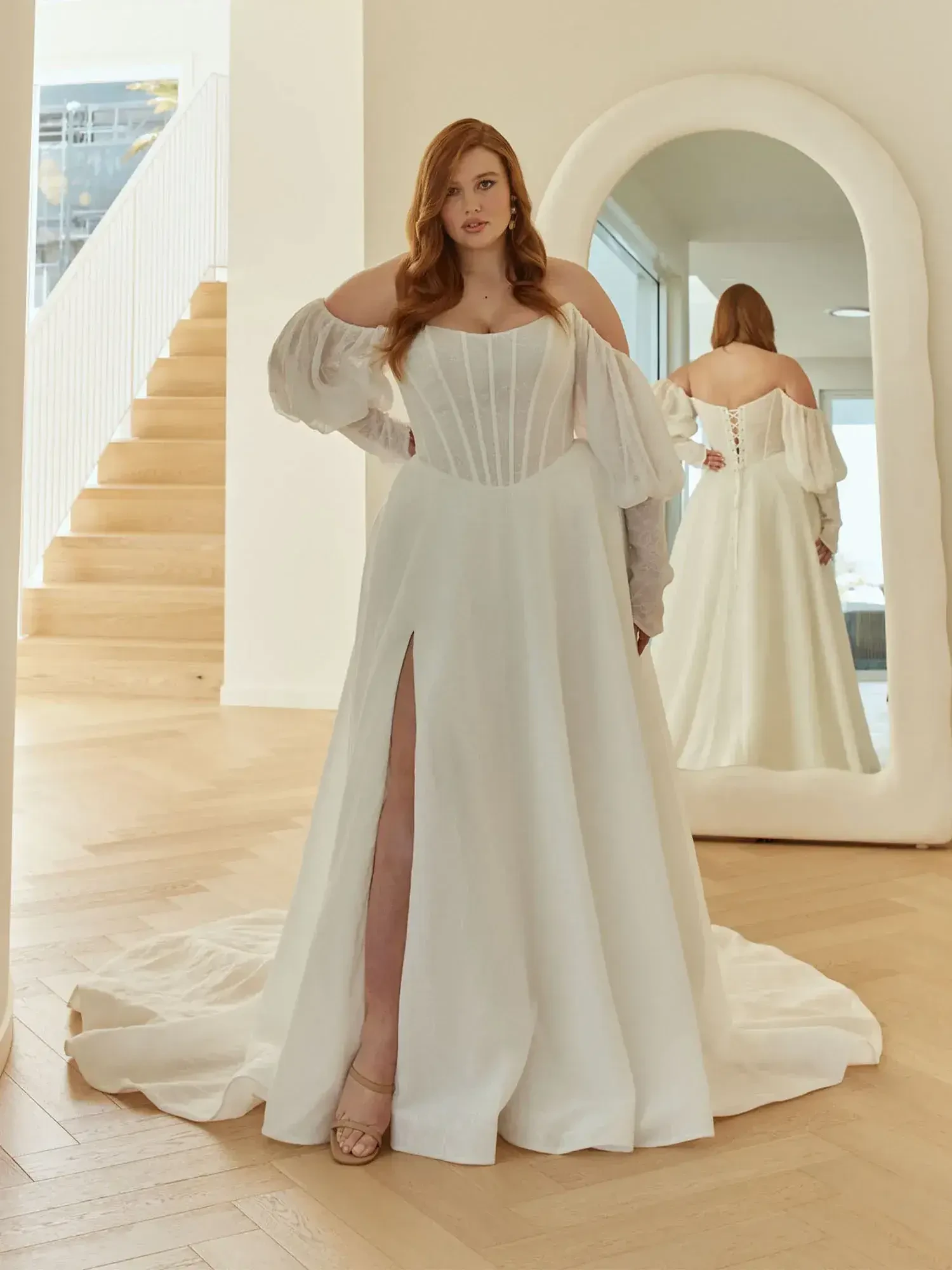 A woman in a flowing white wedding dress with puffed sleeves poses confidently in a sunlit room. A mirror behind her shows the dress's back.