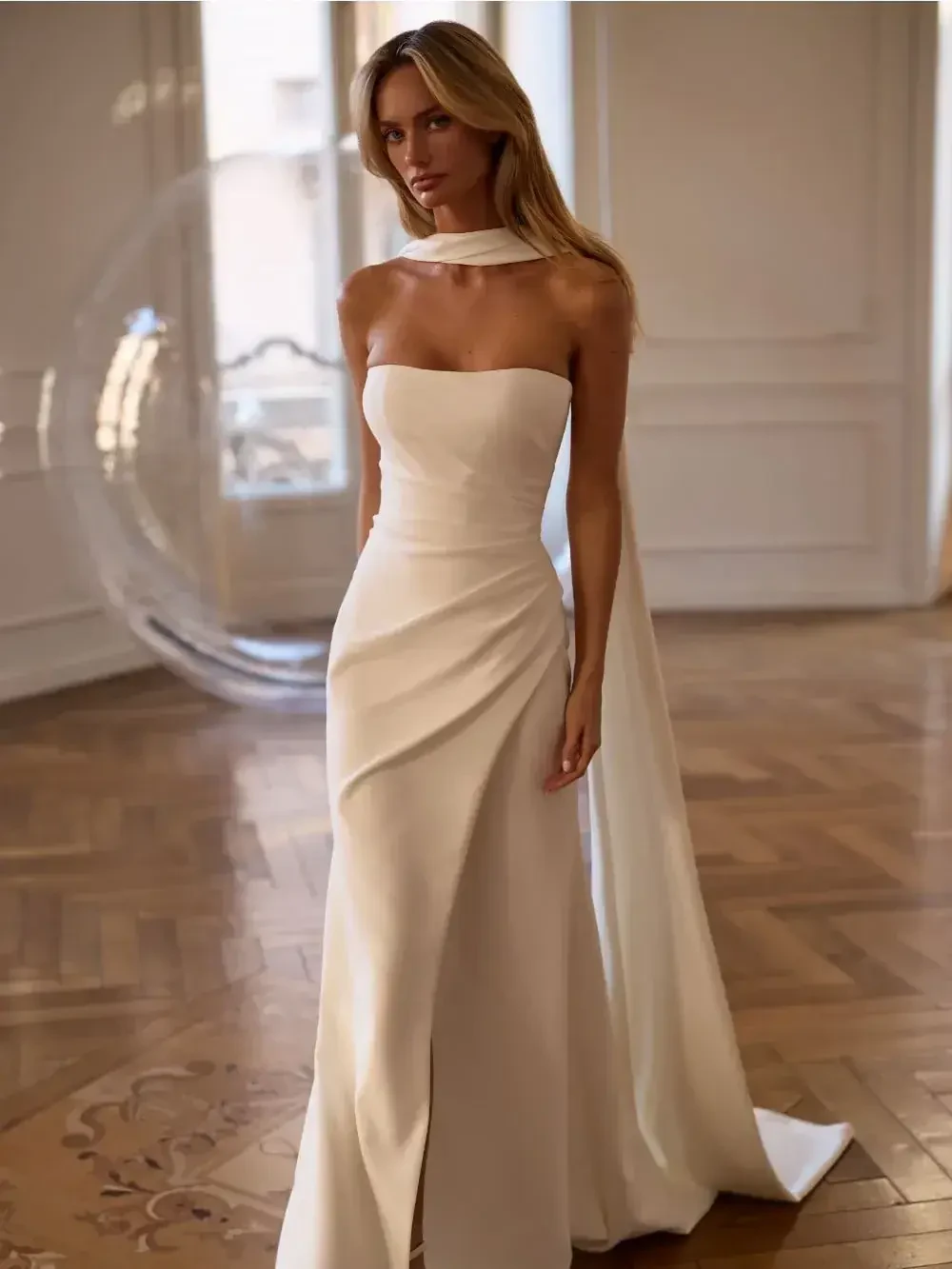 A woman wearing a sleek, strapless white gown with a high slit and a flowing cape in an elegant indoor setting.