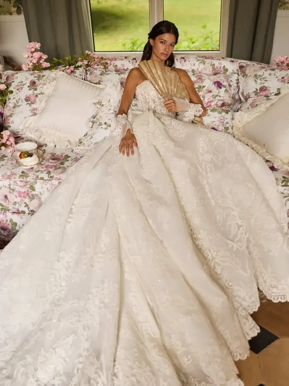 A woman in an elaborate white gown sits on a floral sofa, holding a fan.