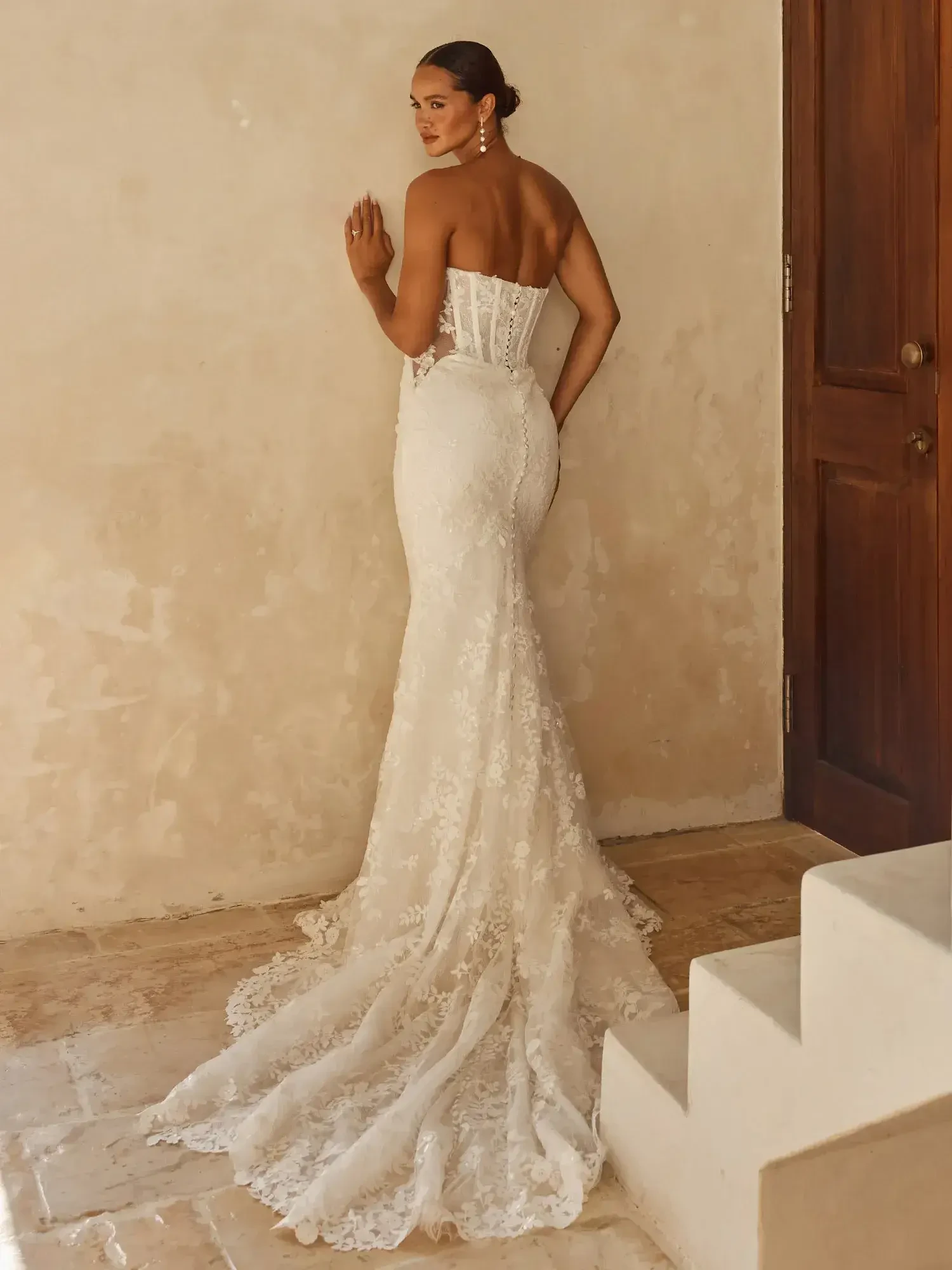 Bride in elegant, strapless lace wedding gown stands by a textured wall and wooden door. The dress features a fitted bodice and flowing train.